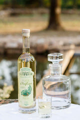 Flasche Fleur d'Angélique Likör neben einer Kristallkaraffe und einem Glas, arrangiert auf weißem Tuch. Im Hintergrund ein Teich.