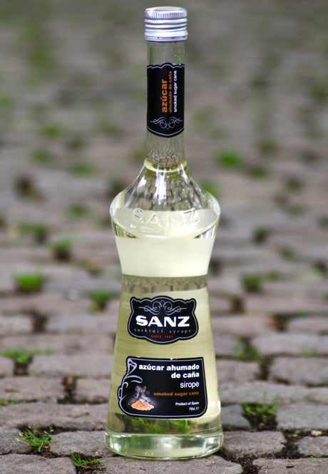 Flasche Sanz geräucherter Rohrzuckersirup auf Kopfsteinpflaster, mit schwarzem Etikett und stilvollem Design.