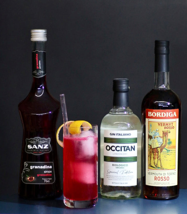 Ginny Cocktail im Highball-Glas mit Zitronenscheibe vor Flaschen von Grenadine Sanz, Occitan Gin und Bordiga Vermouth Rosso.