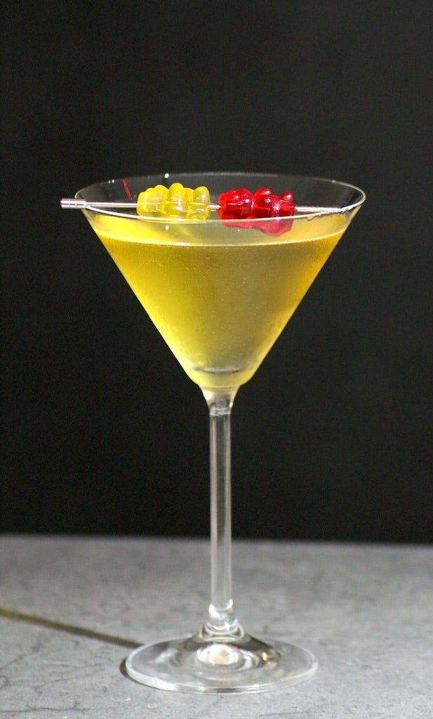 Goldgelber Golden Ermine Cocktail im Martiniglas, garniert mit einem gelben und einem roten Gummibärchen auf einem Spieß.