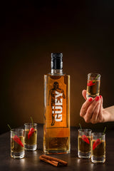 Güey Hot Spicy Gold Tequila, präsentiert mit Chilischoten, Zimtstangen und einem Glas in der Hand vor dunklem Hintergrund.