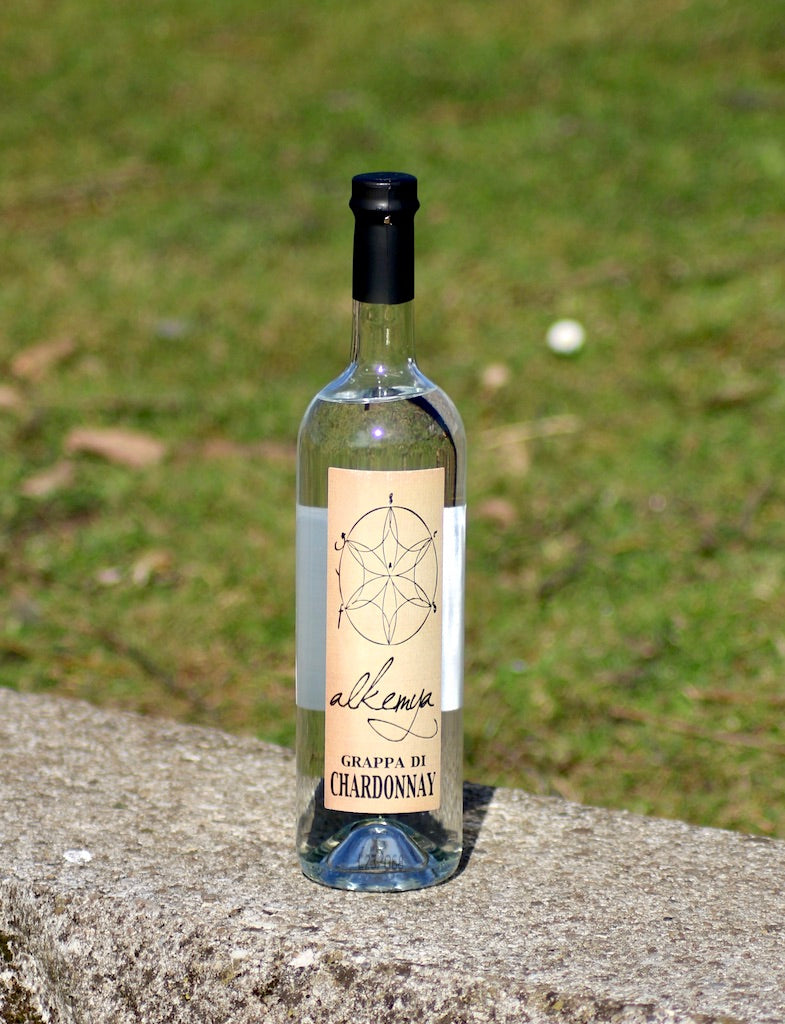 Flasche Grappa di Chardonnay mit hellem Etikett, stehend auf einer Steinmauer vor einer grünen Wiese im Hintergrund