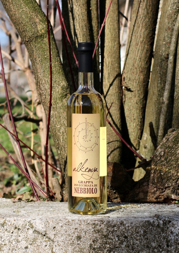 Flasche Grappa Invecchiata di Nebbiolo mit goldfarbenem Etikett, stehend auf einer Steinmauer vor Bäumen.