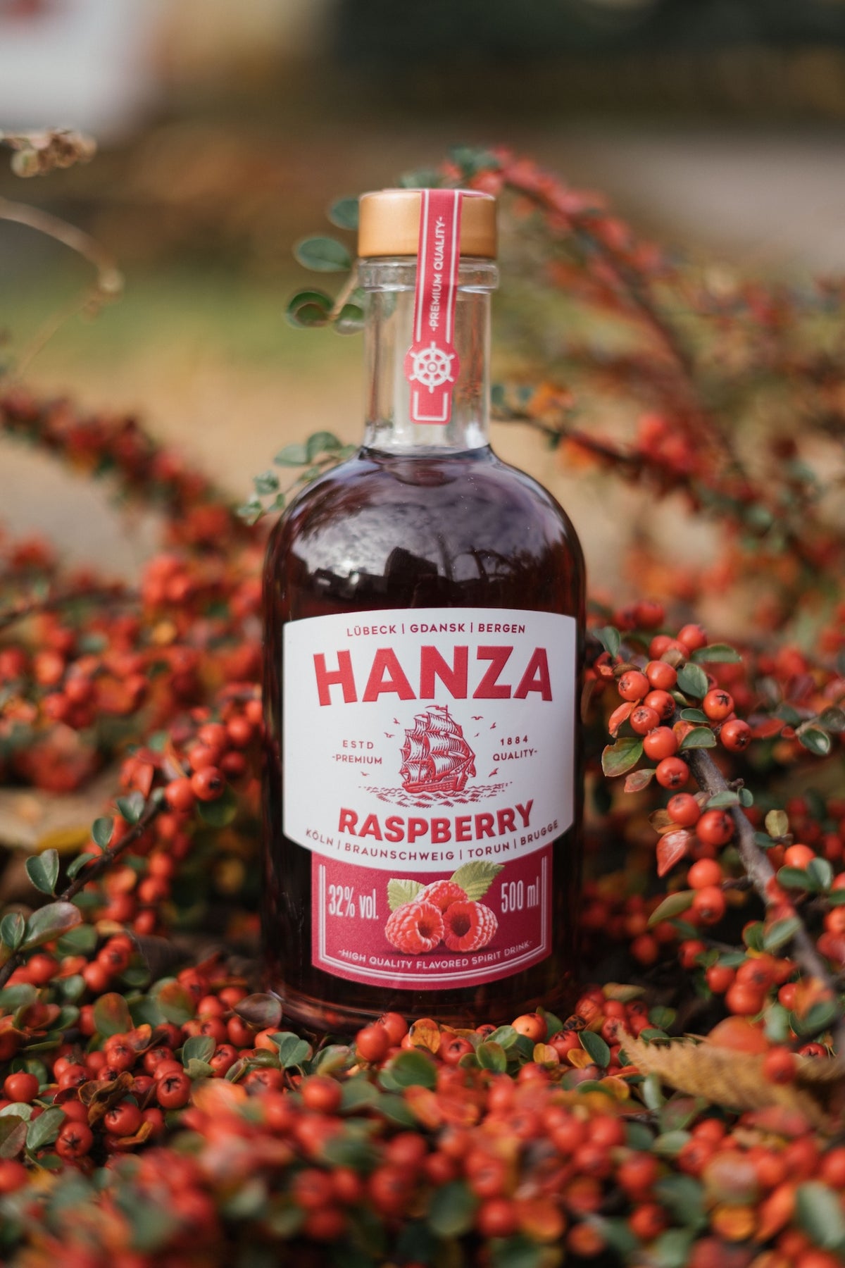 Hanza Raspberry Spirit Drink Flasche umgeben von roten Beeren und Herbstlaub. Rotes Etikett mit Segelschiff-Motiv.