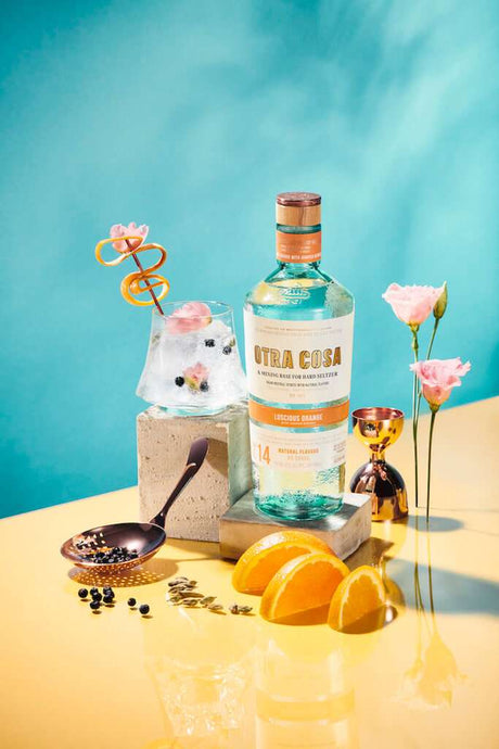 Otra Cosa Luscious Orange Spirituose mit Orangen, Cocktailglas und floralen Akzenten auf farbenfrohem Hintergrund.