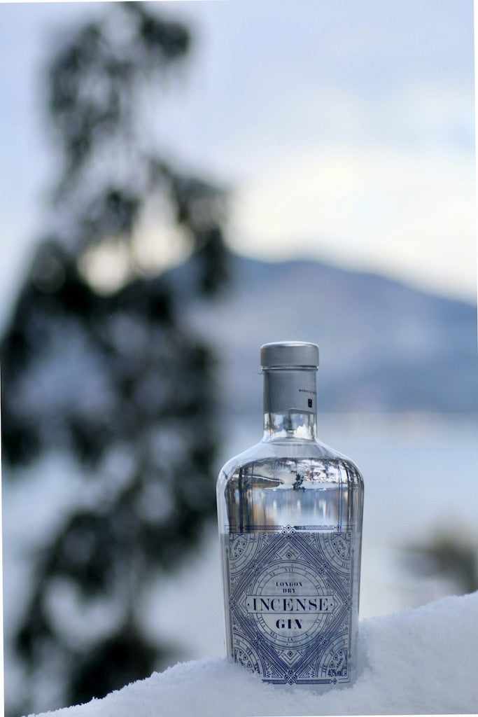 Flasche Incense London Dry Gin mit blauem Etikett, stehend im Schnee vor unscharfem Hintergrund einer winterlichen Landschaft.