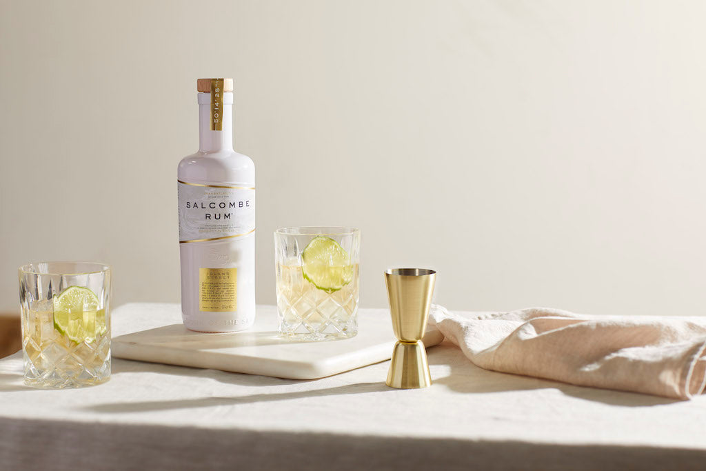 Salcombe Island Street Rumflasche mit zwei Tumblern, Limettenscheiben und Barmaß auf heller Tischfläche.