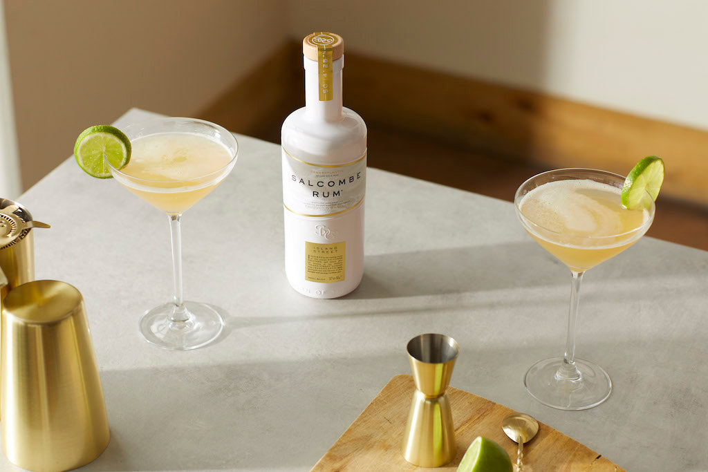 Spiced Pineapple Daiquiri mit Salcombe Rum in Coupettegläsern, Limettenscheiben und Barzubehör auf heller Fläche.