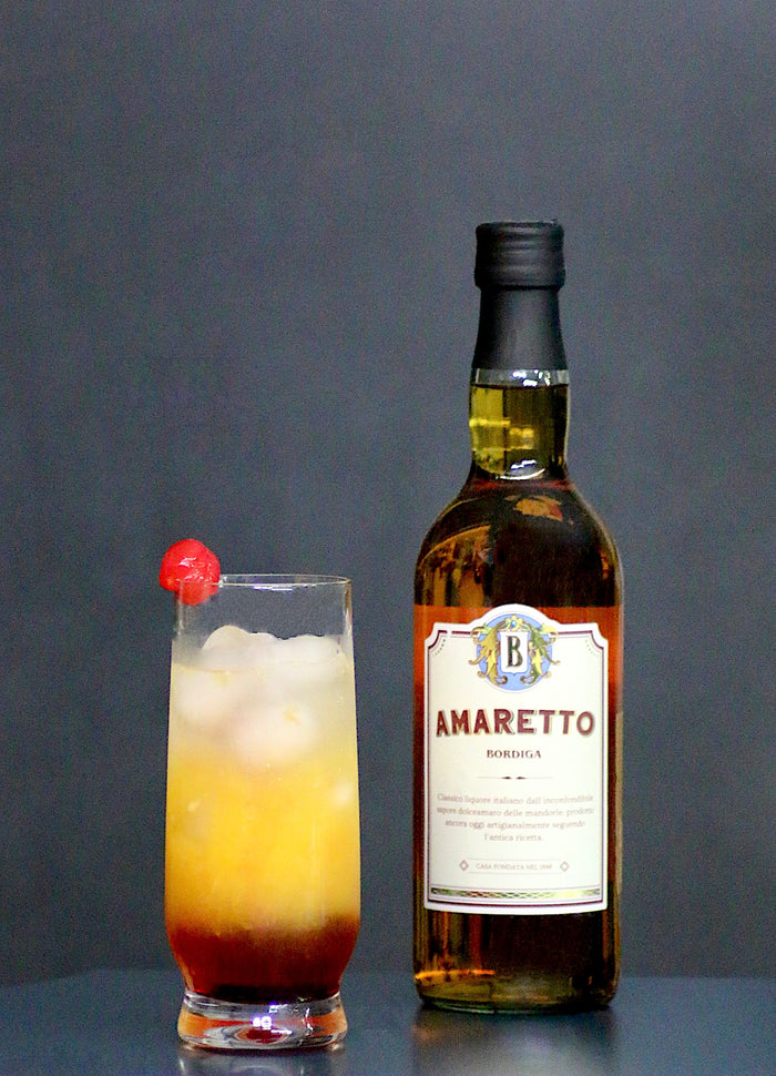Italian Sunset Cocktail im hohen Glas mit Eis und Cocktailkirsche, daneben eine Flasche Amaretto Bordiga vor dunklem Hintergrund.