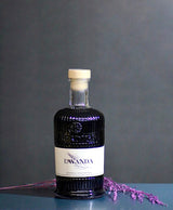Flasche Bordiga Lavanda Likör mit cremefarbenem Etikett und Lavendelzweig auf glänzendem Tisch vor grauem Hintergrund.