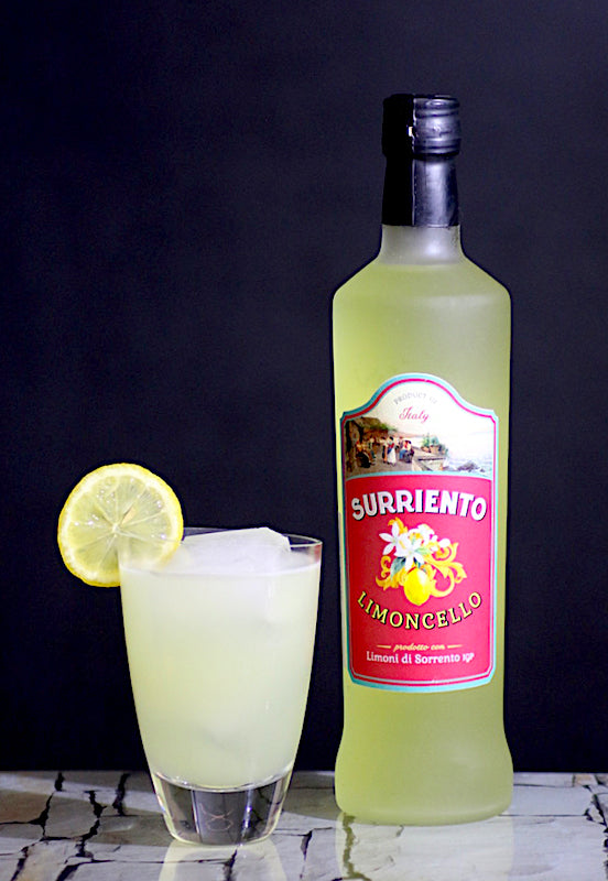 Flasche Limoncello Surriento neben einem trüben, hellgelben Lemon Burst Cocktail mit Eis und Zitronenscheibe vor dunklem Hintergrund.