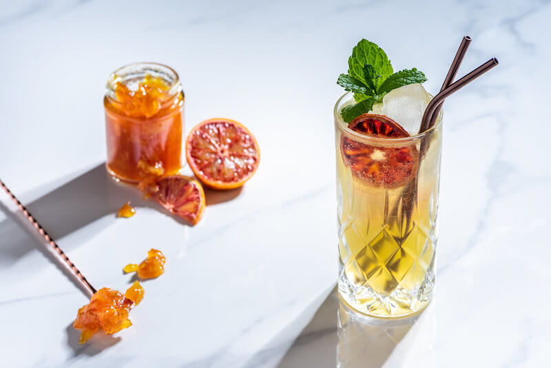 Marmajito Cocktail mit Eiswürfeln im Longdrinkglas, dekoriert mit Minze und getrockneter Blutorange, daneben Marmeladenglas und Löffel.