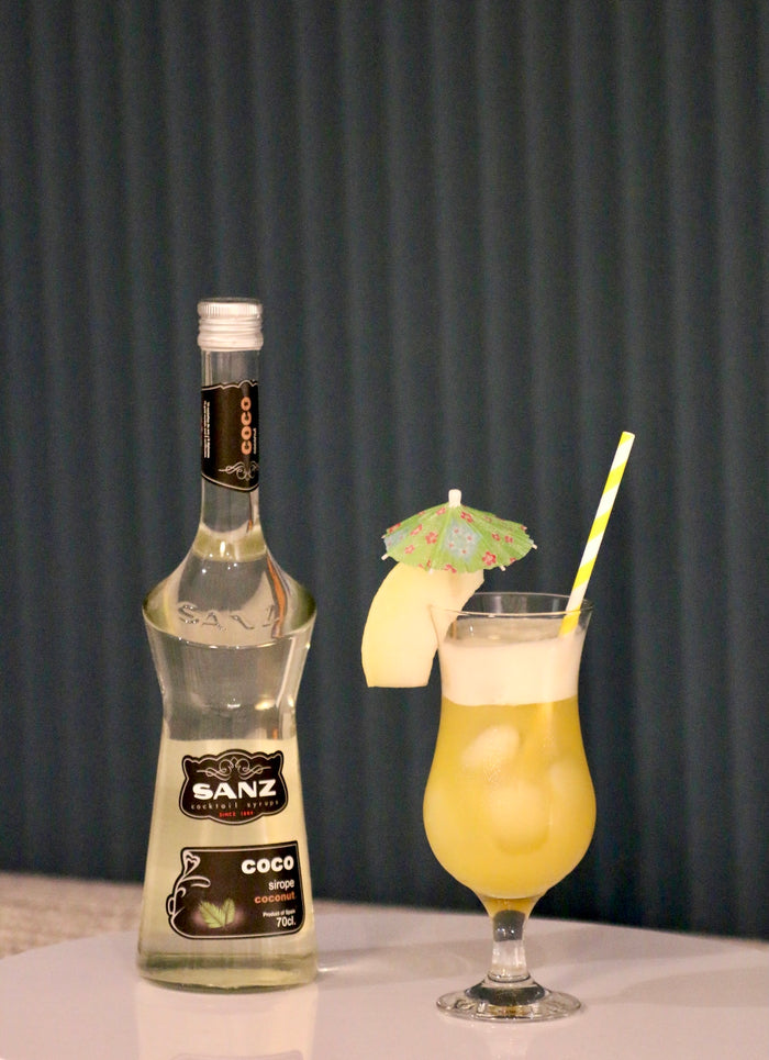 Melon del Sol Cocktail im Hurricane-Glas mit Sanz Kokossirup-Flasche, Melonenscheibe, grünem Schirmchen und gestreiftem Strohhalm.