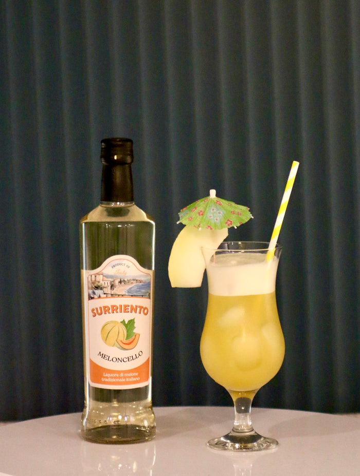 Melon del Sol Cocktail im Hurricane-Glas mit Surriento Meloncello-Flasche, Melonenscheibe, grünem Schirmchen und gestreiftem Strohhalm.