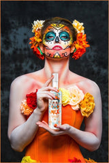 Frau im Catrina-Stil mit farbenfrohem Make-up und Blumen hält eine Flasche Mex Mango-Tequila Likör mit Sahne vor dunklem Hintergrund.
