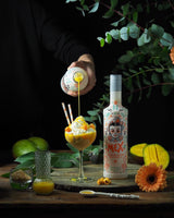 Mex Mango-Tequila Likör mit Sahne, kunstvoll serviert mit Dessert, frischen Mangos und dekorativen Elementen auf rustikalem Holztisch.