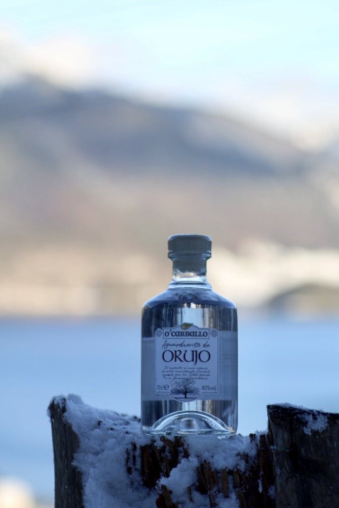 O’Carballo Orujo Flasche im Schnee auf Holzstumpf mit Berg- und Seepanorama im Hintergrund, klarer Tresterschnaps, 70 cl, 40 % vol.