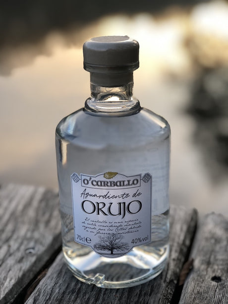 O’Carballo Orujo Flasche auf Holzfläche vor unscharfem Seehintergrund bei Sonnenuntergang, 70 cl, 40 % vol.