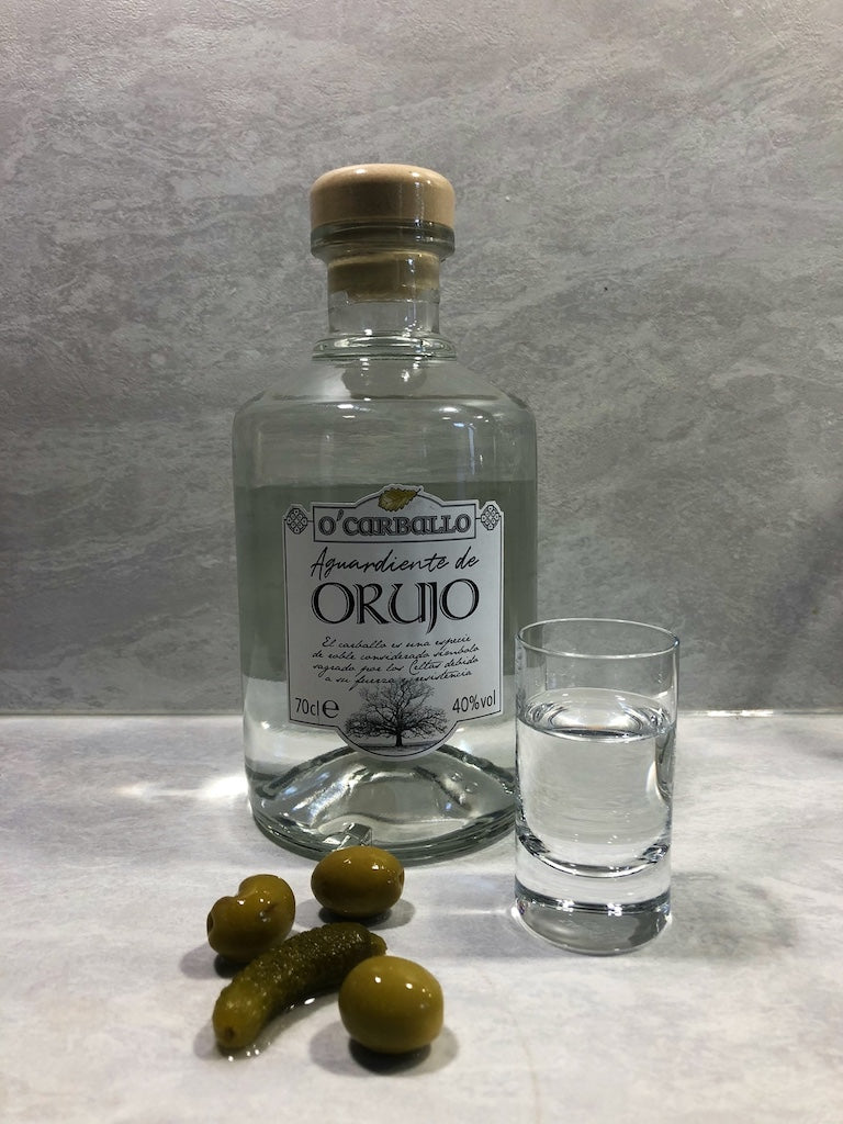 O’Carballo Orujo Flasche mit gefülltem Schnapsglas, grünen Oliven und Cornichon auf hellem Untergrund, 70 cl, 40 % vol.