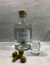 O’Carballo Orujo Flasche mit gefülltem Schnapsglas, grünen Oliven und Cornichon auf hellem Untergrund, 70 cl, 40 % vol.
