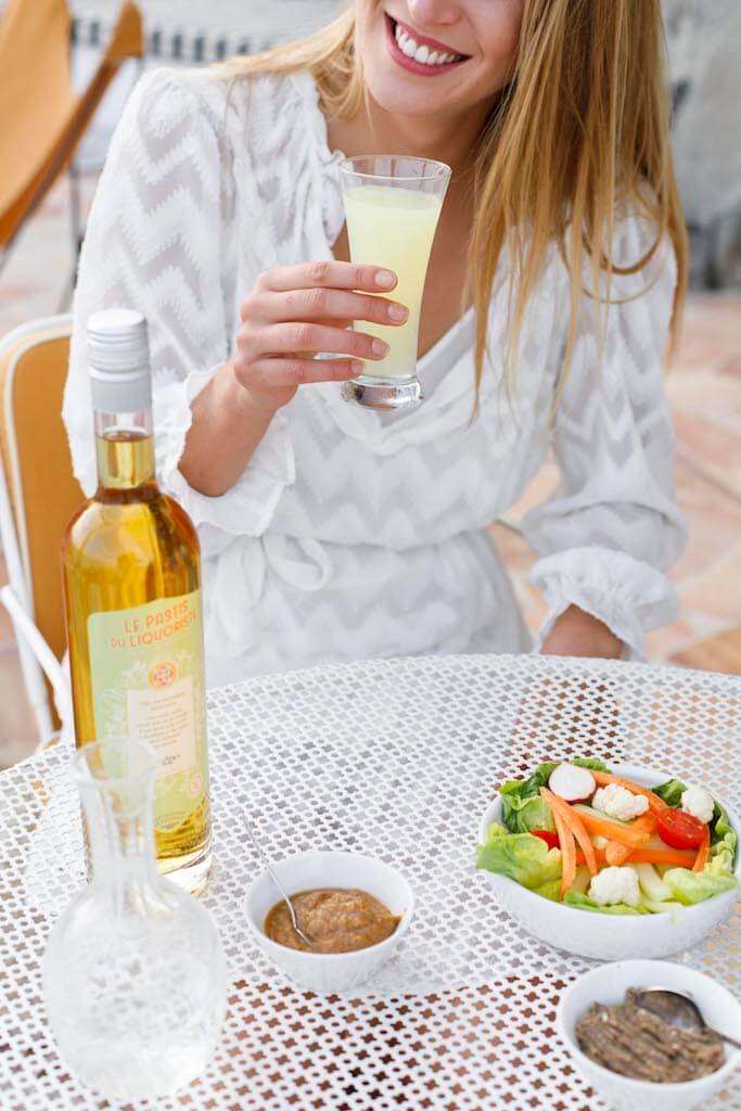 Frau in weißem Kleid hält ein Glas Pastis, auf dem Tisch eine Flasche Pastis du Liquoriste, Karaffe, Salat und Dipschälchen.