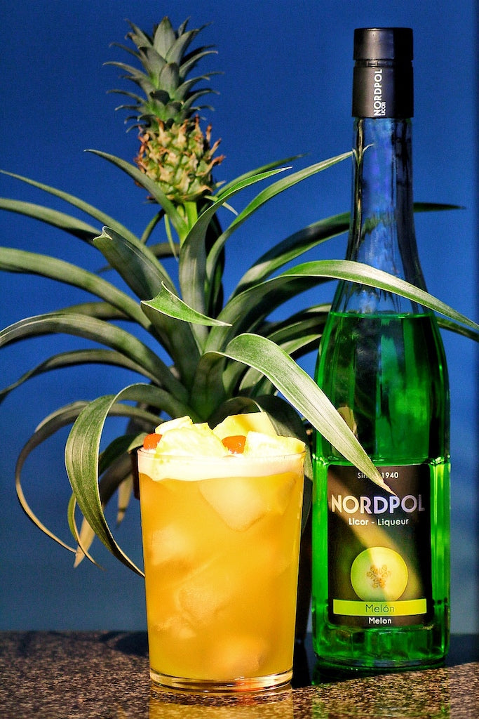 Pearl Harbor Cocktail im Highball-Glas auf Eis mit Ananasstücken, daneben grüne Flasche Melonenlikör und Ananaspflanze im Hintergrund.
