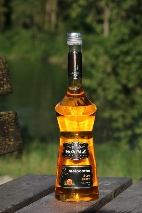 Sanz Pfirsichsirup in bernsteinfarbener Flasche auf Holzbank vor grünem Hintergrund