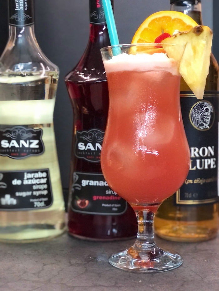 Planter’s Punch Cocktail mit Orangenscheibe, Kirsche und Ananas, daneben Rum, Zuckersirup und Grenadine von Sanz.