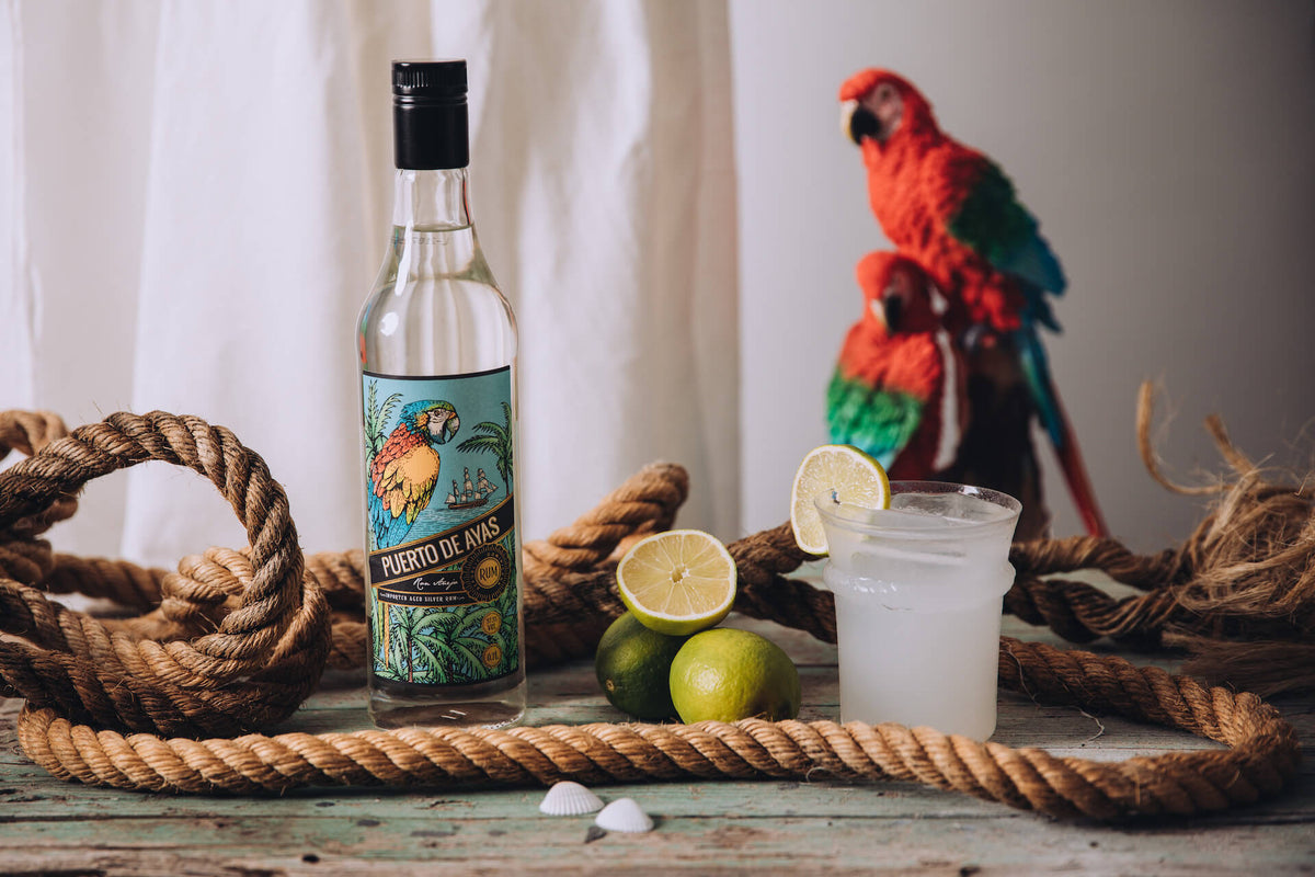 Puerto de Ayas Rumflasche mit Limetten, Seil und Cocktail, im Hintergrund zwei Papageien vor heller Stoffkulisse.