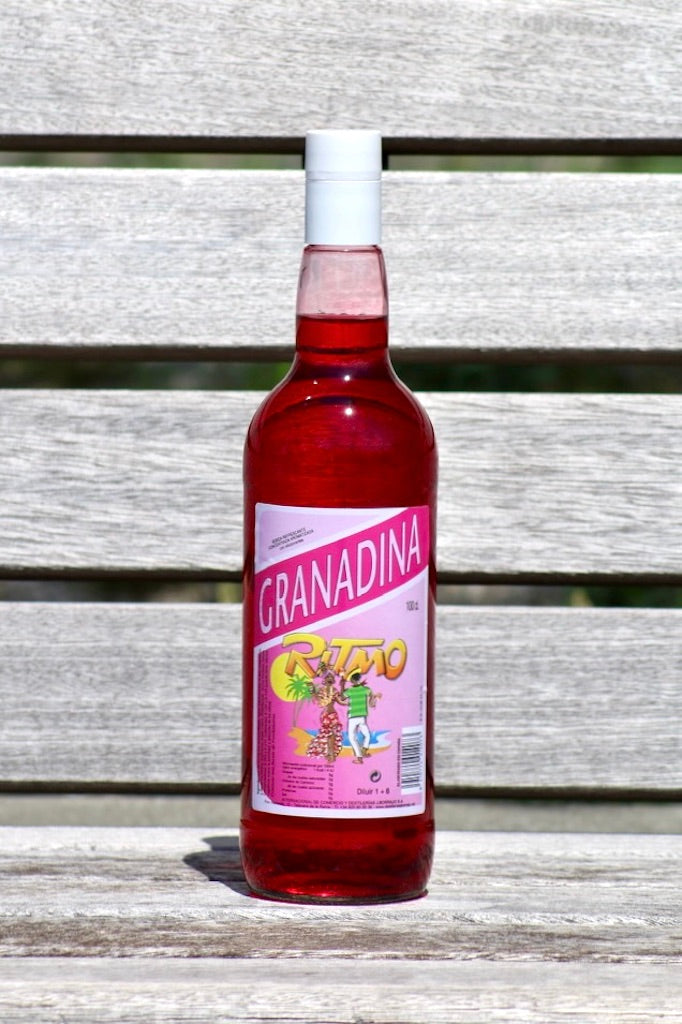 Flasche Ritmo Granadina zuckerfrei, 1 L, mit rosa Etikett und Tanzszene, roter Sirup, weißer Schraubverschluss, auf Holzbank.