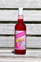 Flasche Ritmo Granadina zuckerfrei, 1 L, mit rosa Etikett und Tanzszene, roter Sirup, weißer Schraubverschluss, auf Holzbank.