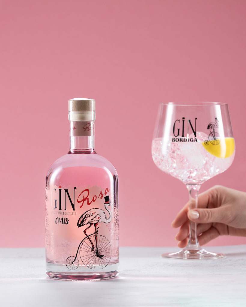 Flasche Ciais Gin Rosa von Bordiga vor rosa Hintergrund, daneben ein Gin Tonic im Ballonglas mit Zitronenzeste, gehalten von einer Hand.