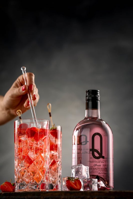 Rose Gin & Tonic beim Mixen mit Rose Gin Q Flasche, Eis und Erdbeeren vor dunklem Hintergrund