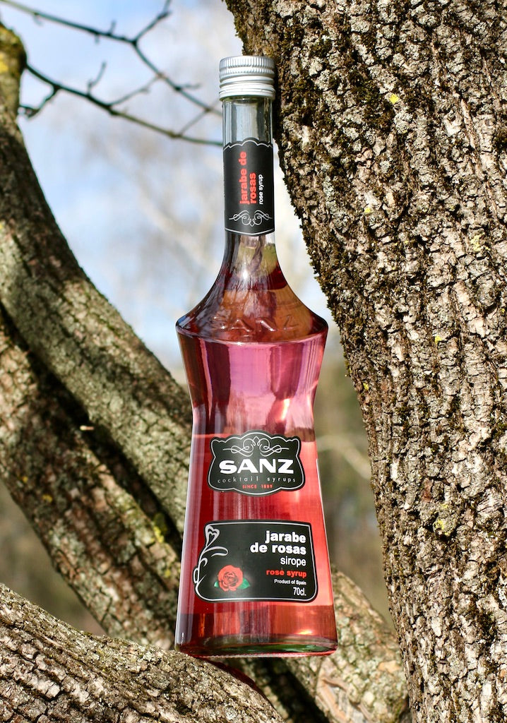 Sanz Rosensirup 700 ml, elegante Flasche mit schwarzem Etikett, präsentiert vor einer Baumrinde mit natürlichem Hintergrund.