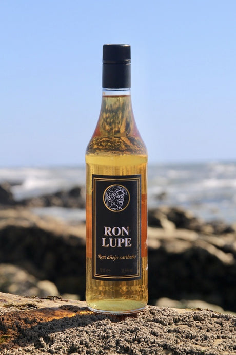 Rum Añejo Lupe, goldbraune Flasche mit schwarzem Etikett, am Meer auf Felsen platziert mit blauem Himmel im Hintergrund.