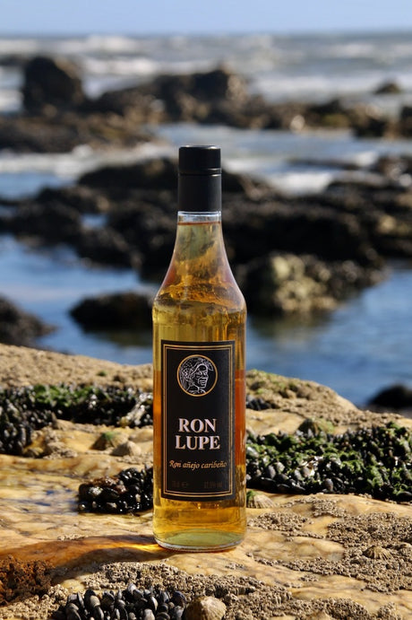 Rum Añejo Lupe mit schwarzem Etikett, goldbraune Flasche auf Felsen am Meer mit Wellen im Hintergrund.
