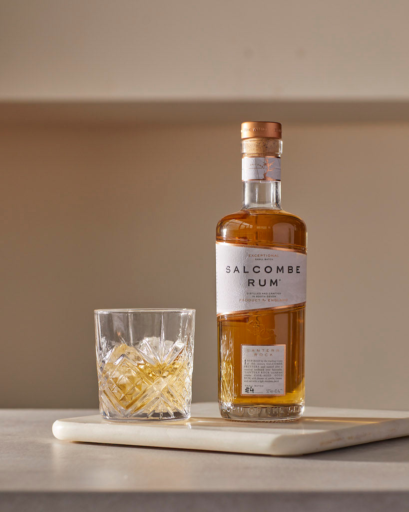 Flasche Salcombe Rum und ein Tumbler-Glas mit Eis auf einer hellen Oberfläche.