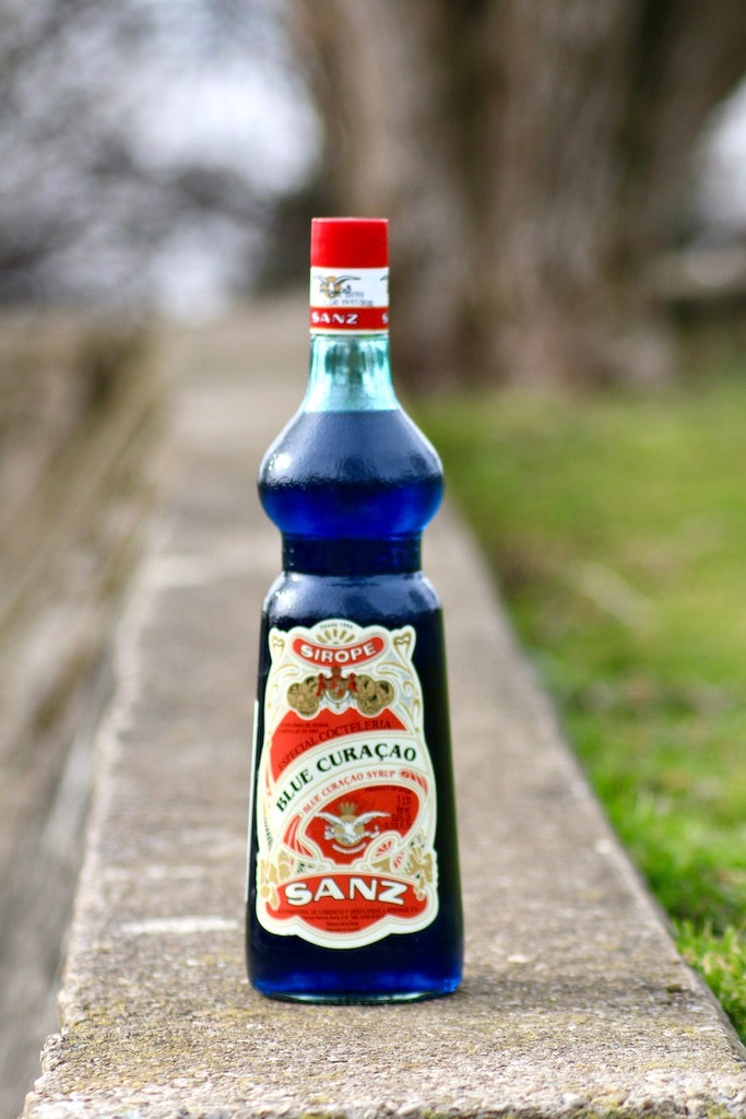 Flasche Sanz Blue Curacao Sirup mit rotem Etikett und Verschluss, 1 Liter, auf einer Steinfläche platziert.