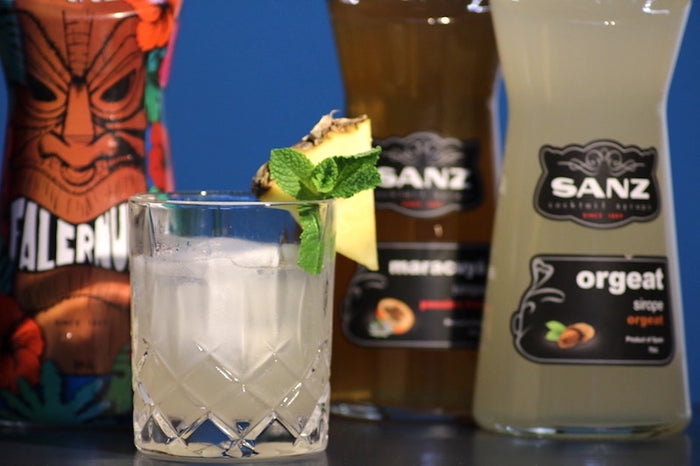 Saturn Cocktail in Kristallglas mit Ananasscheibe und Minze, daneben Samara Gin und Orgeat Sirup von Sanz vor blauem Hintergrund.