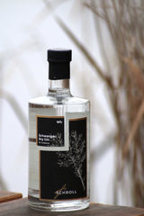Schwangau Dry Gin Flasche mit schwarzem Etikett steht auf einer Holzfläche, im Hintergrund natürliche Gräser und ein heller Himmel.