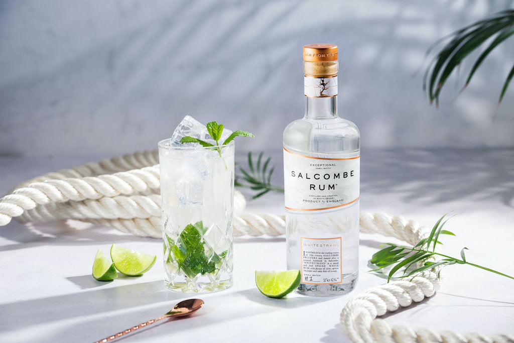 Salcombe Spiced White Rum Whitestrand neben einem Mojito mit Minze und Limetten, präsentiert auf einem weißen Hintergrund mit maritimen Seilen.