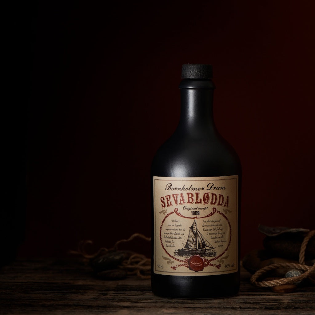 Bornholmer Dram Sevablødda in dunkler Flasche mit maritimem Etikett, rustikal inszeniert mit Seilen und Natursteinen.