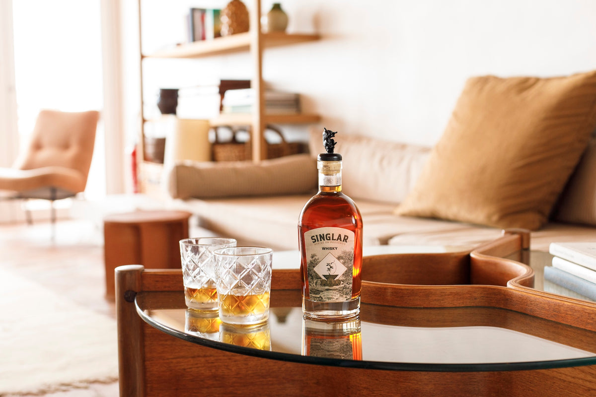 Flasche Singlar Whisky auf einem Glastisch, daneben zwei gefüllte Gläser, im Hintergrund ein Sofa mit Kissen.