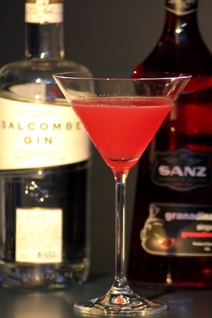 Roter Cocktail aus Gin und Grenadine in Martini-Glas mit Flaschen im Hintergrund