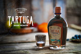 Tartuga Rumflasche mit Glas auf Holztisch, unscharfer Hintergrund mit Obst und Schriftzug Tartuga