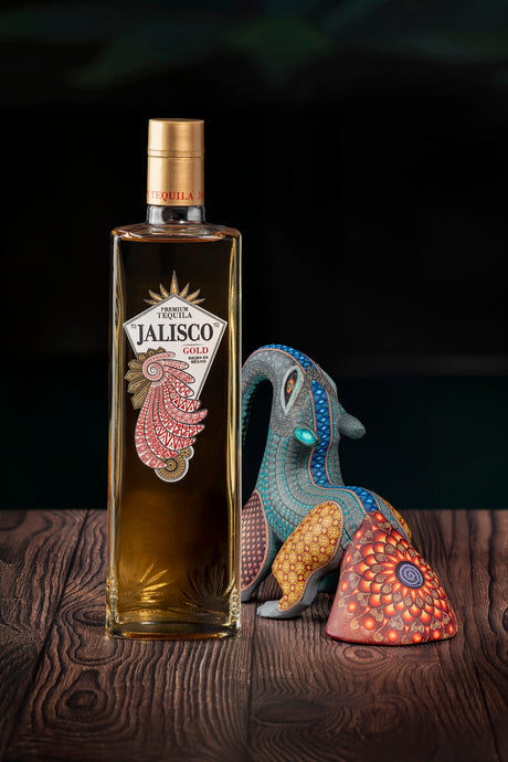 Tequila-Jalisco-Gold-Flasche auf Holzoberfläche neben einer bunt bemalten mexikanischen Alebrije-Figur.