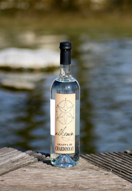 Flasche Grappa di Chardonnay mit hellem Etikett, stehend auf einem Holzsteg vor einem unscharfen Gewässerhintergrund.