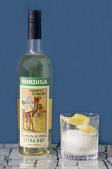 Flasche Bordiga Vermouth di Torino Extra Dry mit Kamelmotiv, neben Glas mit Eis und Zitronenschale vor blauem Hintergrund.