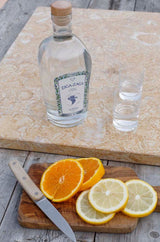 Flasche Ziga Zaga Vodka mit zwei Gläsern auf Steinplatte, davor Orangen- und Zitronenscheiben mit Messer auf Holzbrett.