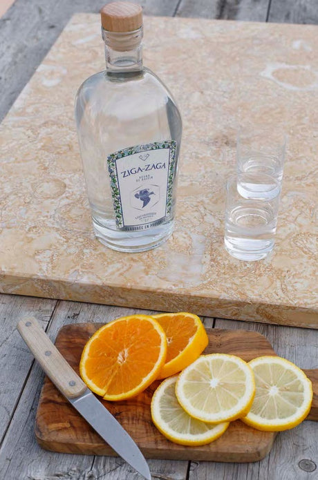Flasche Ziga Zaga Vodka mit zwei Gläsern auf Steinplatte, davor Orangen- und Zitronenscheiben mit Messer auf Holzbrett.